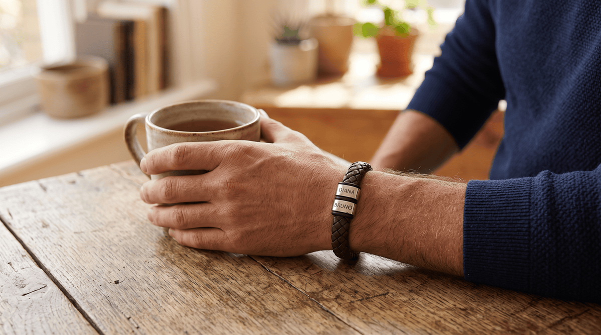 A close-up of a man's hands resting on a rustic wooden table, holding a ceramic mug. He is wearing a dark brown braided leather bracelet with silver-toned metal nameplates engraved with 'JOHN' and 'DEAR'.