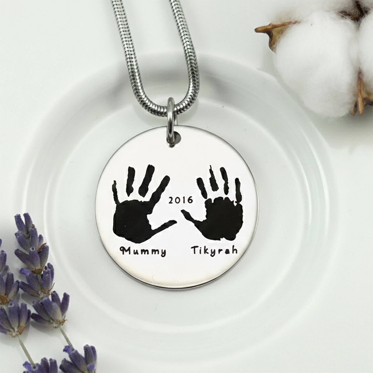 Belle Fever Hand on My Heart Necklace — silver round pendant engraved with handprints, displayed on a soft surface, a meaningful keepsake symbolising love and remembrance.