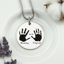 Belle Fever Hand on My Heart Necklace — silver round pendant engraved with handprints, displayed on a soft surface, a meaningful keepsake symbolising love and remembrance.